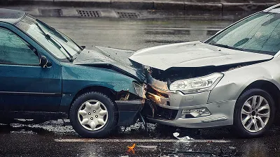 A head-on collision between a dark teal car and a silver car, showing significant front-end damage and crumpled hoods on both vehicles.