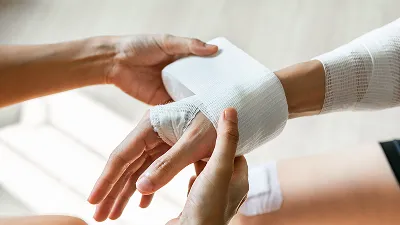 A person's hands carefully wrapping a white gauze compression bandage around another person's wrist and palm.