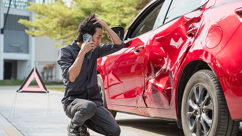 A man is kneeling down next to a red car that has been in an accident.